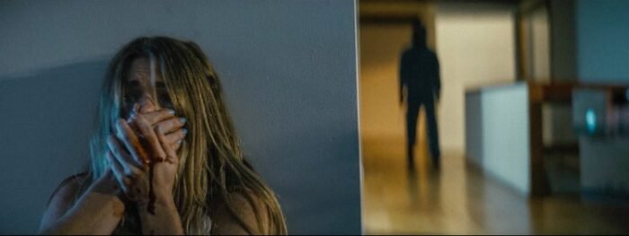 A woman hides behind a wall holding her mouth shut in her blood-dripping hands as a dark figure stalks her in the background.