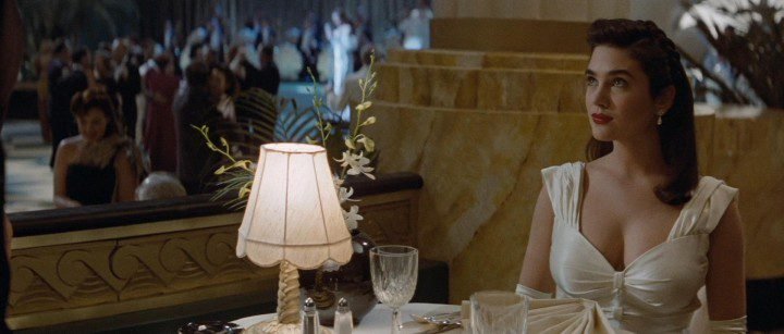 A woman with long dark hair sits at a table looking off camera, she is wearing a white gown with a plunging neckline