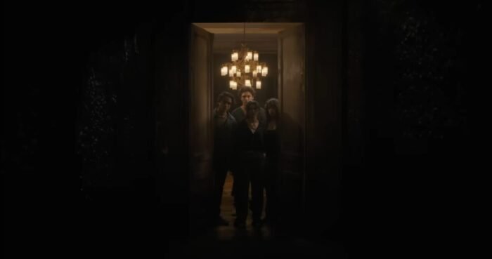 a group of young men and women standing at the threshold of a giant door, a chandelier hangs above them in the background.