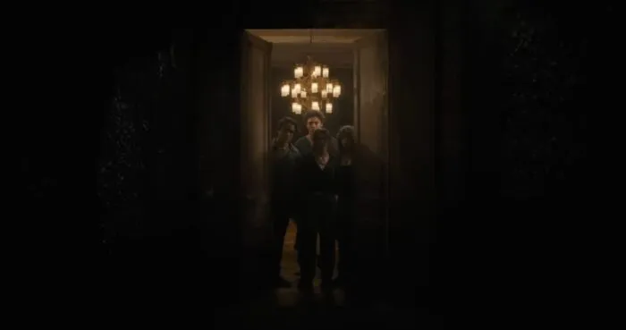 a group of young men and women standing at the threshold of a giant door, a chandelier hangs above them in the background.