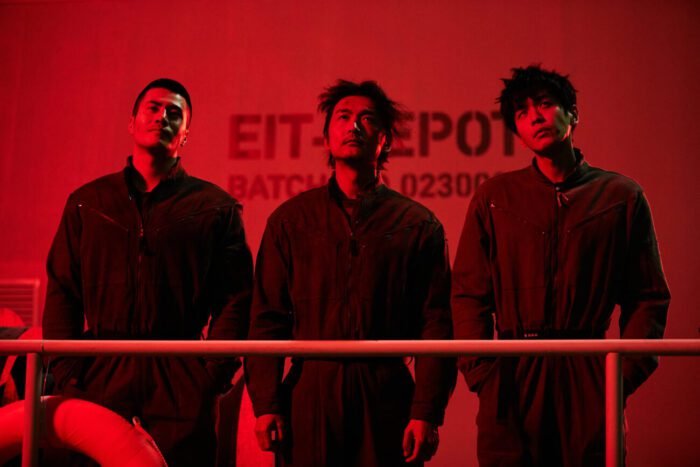 Three men are seen behind a railing under a red light in ESCAPE FROM THE 21ST CENTURY