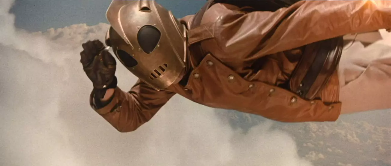 A man in a leather jacket and bronze helmet salutes at the camera as he flies with clouds behind him