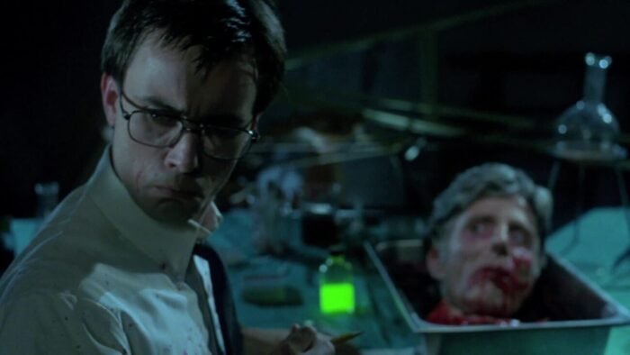 A decapitated head rests on a metal tray beside a glowing green vile and various medical instruments while a man in the foreground looks over his shoulder.
