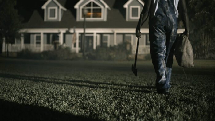 a man in overalls is seeing walking across a lawn toward a house with an ax in one hand and a burlap sack in the other.
