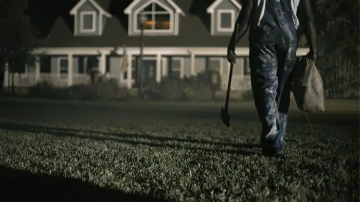 a man in overalls is seeing walking across a lawn toward a house with an ax in one hand and a burlap sack in the other.