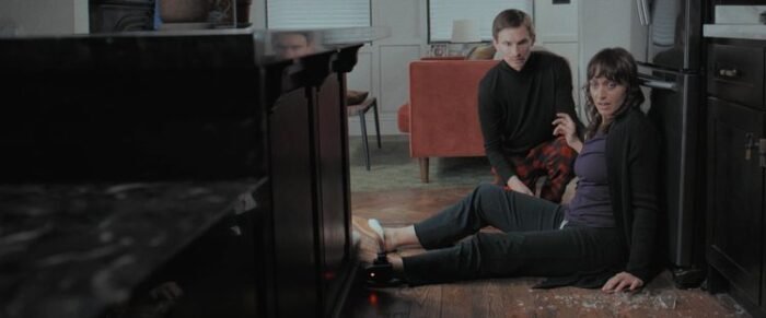 A woman sits on the floor with her back to the wall, scared about something toward the camera she's gesturing toward as the man kneeling beside her attempts to console her.