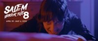 A woman is seen at the foot of a bed bowing her head while looking toward the camera with an evil smile in HOUSE OF ASHES