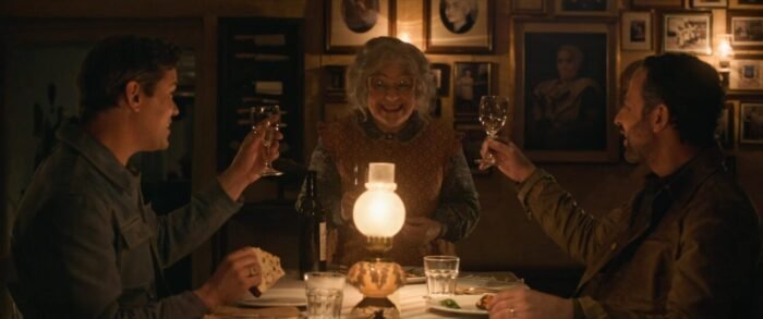 Two men sitting at a table lit by candlelight , raise their glasses to the woman standing at the head of the table.