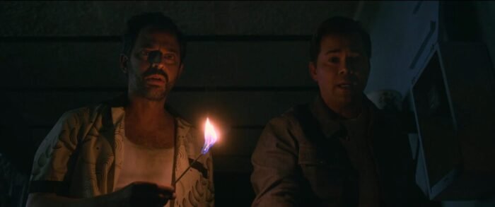Two men look worried, lit by a match one is holding.