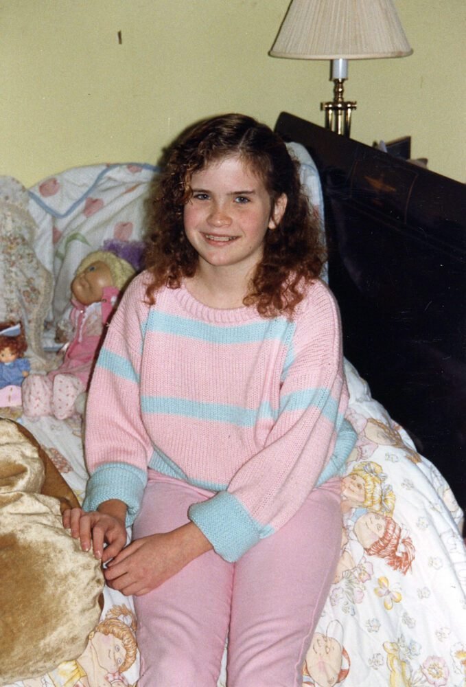 a young girl smiles for the camera while sitting on the edge of a bed.