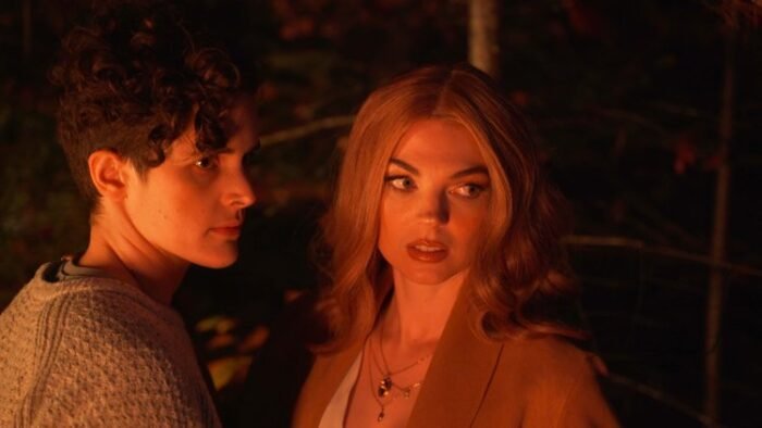 Two women, lit by the light of a nearby campfire, shoot a look at someone off camera.