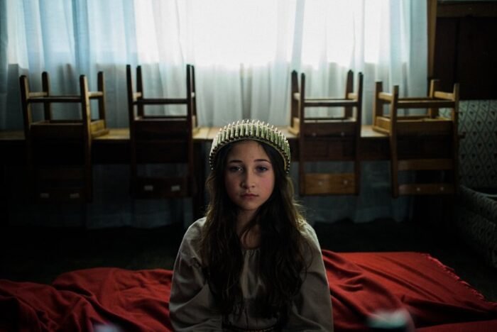 A young girl in a crown of ammunition sits on the edge of a bed. behind her a row of chairs are upside down on furniture against the window.