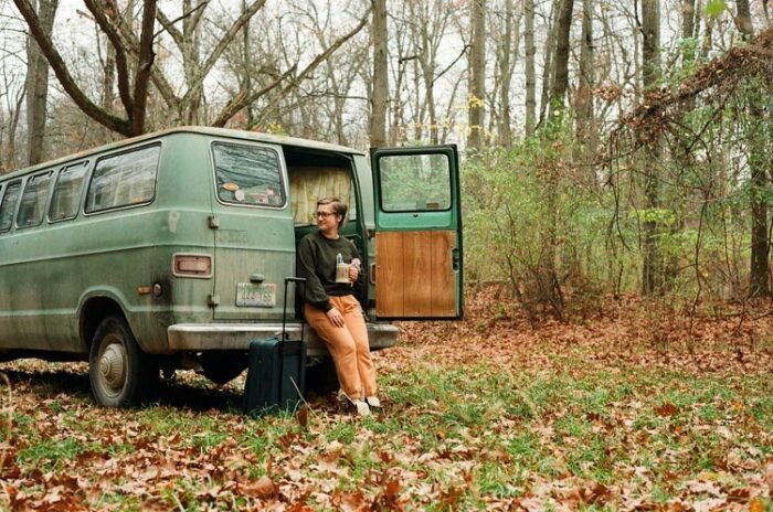 A woman sits against her van's rear bumper in a heavily wooded area.