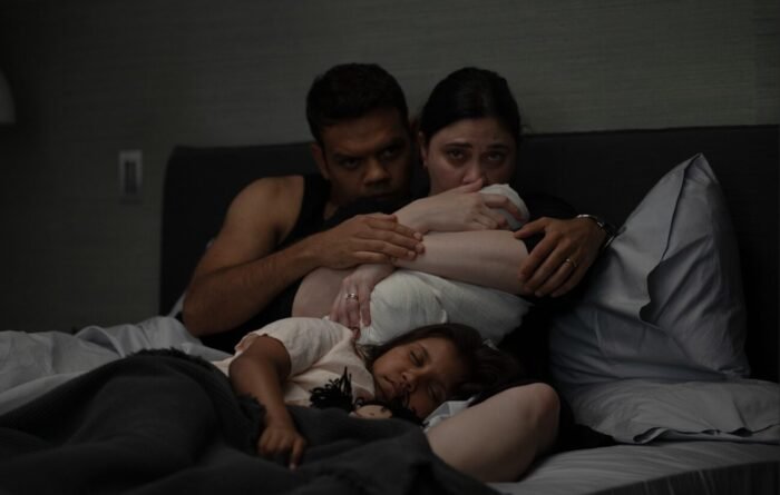 A mother holds her baby while her husband holds her and their daughter sleeps against in them in their bed.