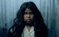 A child with dark, flowing hair is seen expressionless with white eyes.