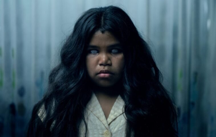 A child with dark, flowing hair is seen expressionless with white eyes.