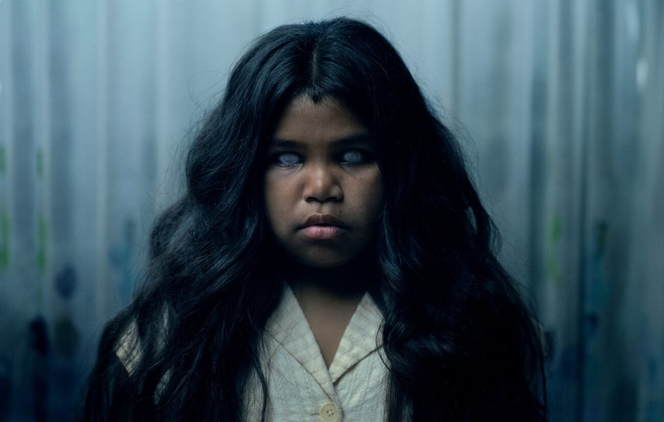 A child with dark, flowing hair is seen expressionless with white eyes.