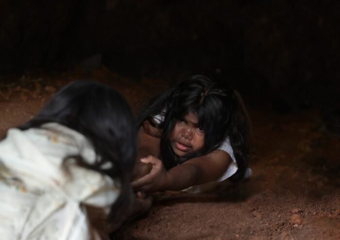 A child being pulled into a cave stretches her arms out to her friend for help in THE MOOGAI
