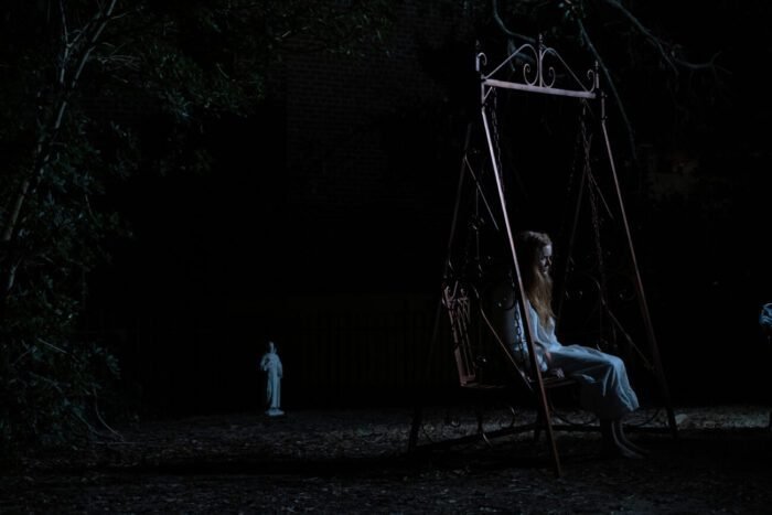 A woman sits on a swing in the dead of night, another figure is standing near the tree line in the background.
