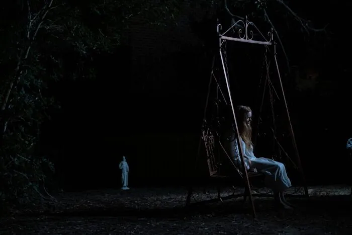 A woman sits on a swing in the dead of night, another figure is standing near the tree line in the background.