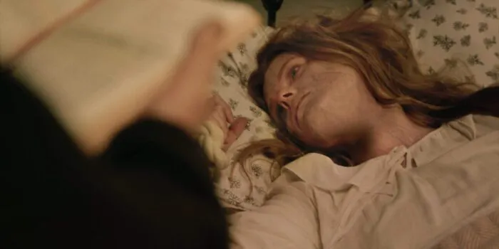 A sick looking woman lies in bed, a pair of hands holding a book n the foreground suggests someone is reading to her.