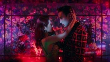 A man and woman embrace in front of a fish tank under neon red and pink lights.