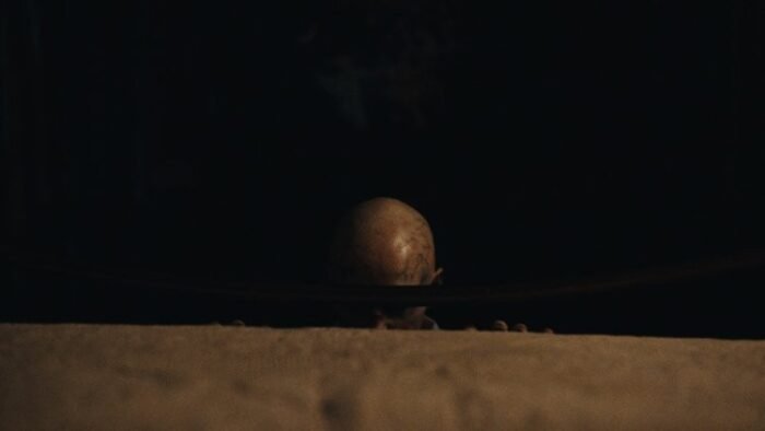 A bald figure crouches at the base of a bed, his eyes can't be seen through the bed frame's cross bar.
