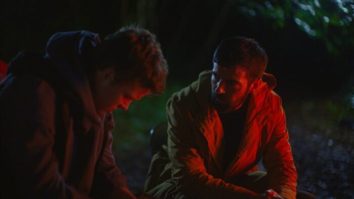 Two men are talking around a campfire that is lighting their faces. (AMONGST THE WOLVES)