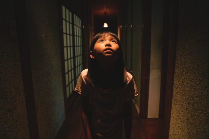 a young girl looks up under a light in a hallway.