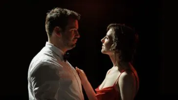 A man and a woman standing together under a spotlight looking in each other's eyes (DO I KNOW YOU FROM SOMEWHERE?)