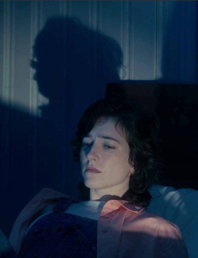 A woman sitting up in bed, her shadow looms on the wall behind her (DO I KNOW YOU FROM SOMEWHERE?)