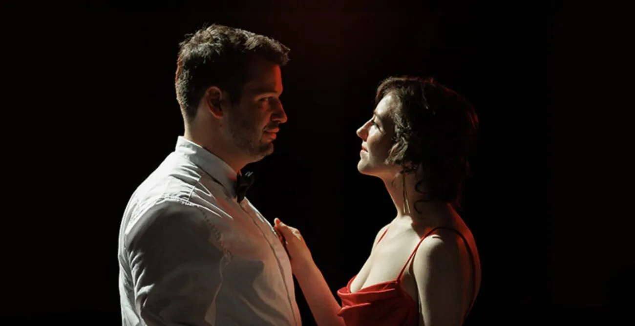 A man and a woman standing together under a spotlight looking in each other's eyes (DO I KNOW YOU FROM SOMEWHERE?)