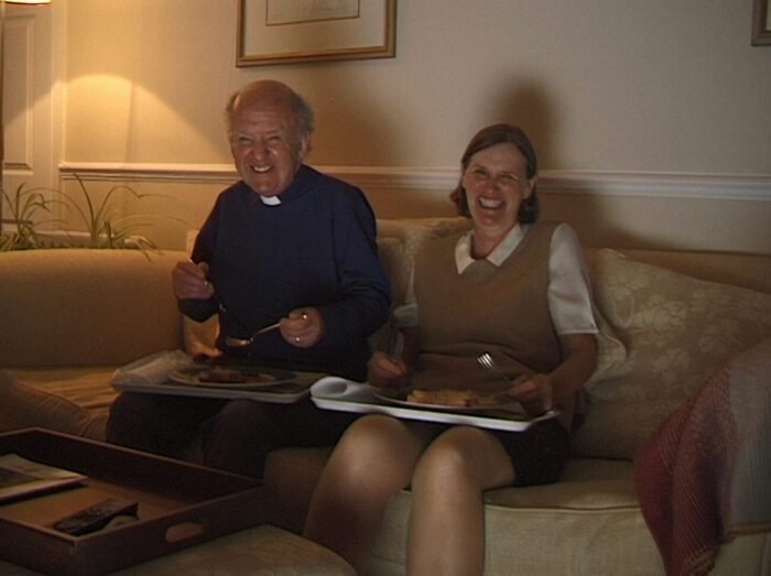 A priest and a woman are seen eating from dinner trays on a living room couch in Four Evil Deeds