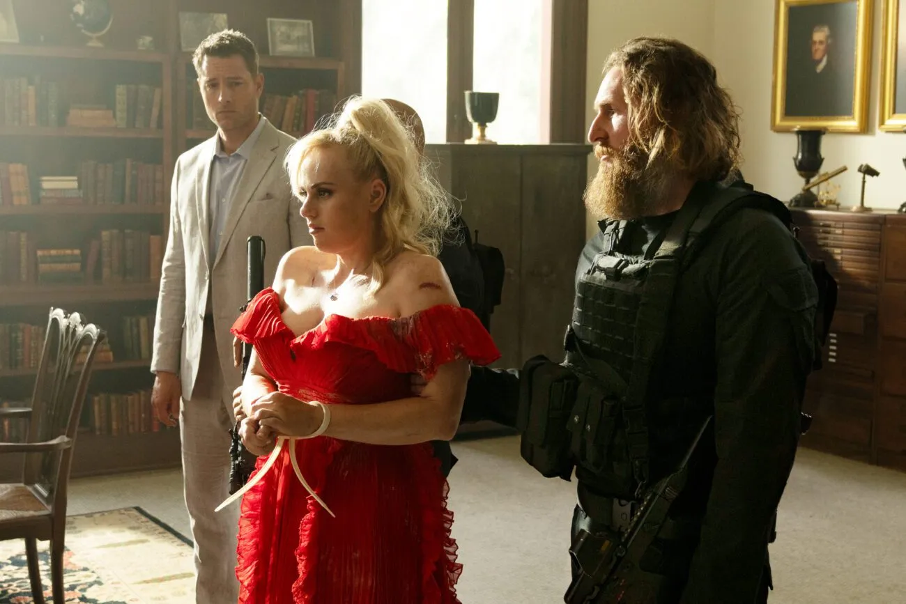 A woman in a red dress is being led by a mercenary with long hair, while a man in a gray suit stands in the background