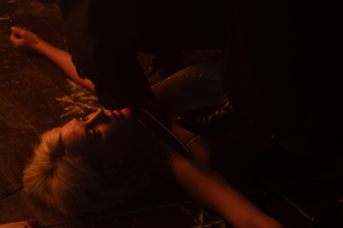 A cloaked figure is holding a knife to the neck of a woman lying on the floor (Hell Motel)
