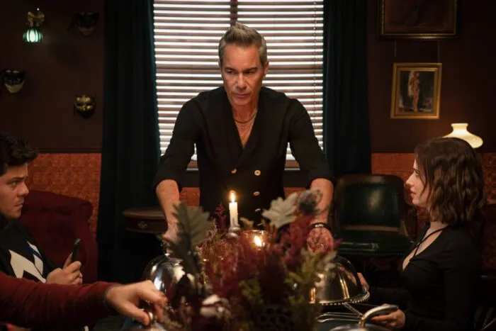 A man lifts a platter onto a dining table where others are sitting. What's on the platter is hidden behind a floral centerpiece (HELL MOTEL)