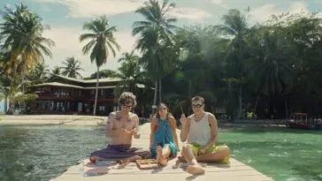 Three people sitting on a dock with a residence on the beach in the background.