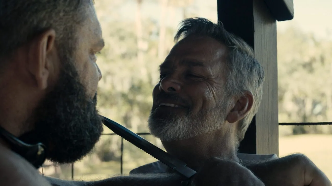 A large bearded man holds a knife to another man's neck.