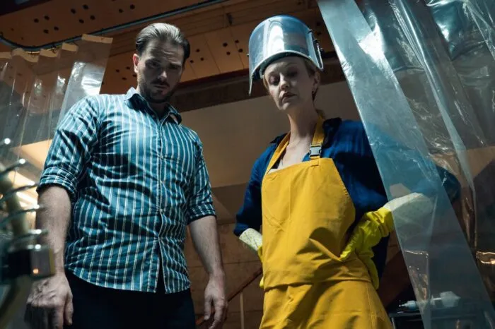 A man in a button-up shirt stands beside a woman in a rubber apron and facemask in Pins and Needles.