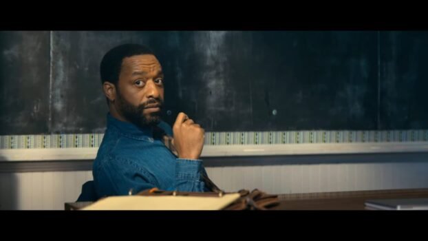 Marty looks at the camera while he's sitting in a chair in front of the classroom blackboard
