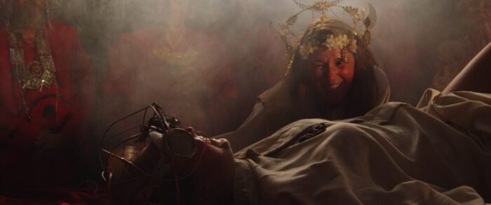A woman in a flower headdress looms over a robed woman tied to a table.