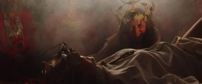 A woman in a flower headdress looms over a robed woman tied to a table.