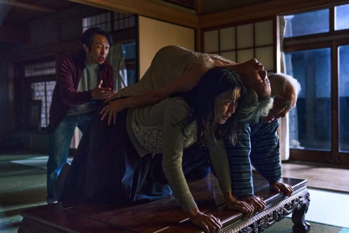 two people create a table for a woman to rest upon their backs, while a man in the back coaches them.