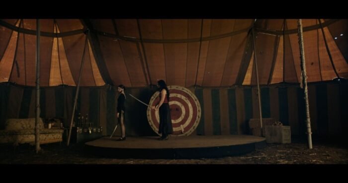 A woman holds a store up to another's back in a circus tent with a bullseye behind them.