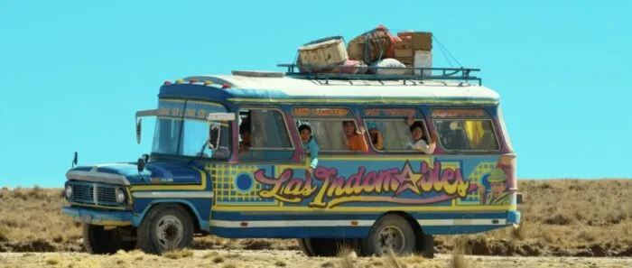 A colorful bus with women hanging out the windows drives through the Bolivian desert