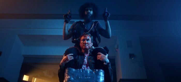 A man sits atop another mans shoulders. He is holding guns in each hand and duct taped to the man he is on top of. (Hold the Fort)