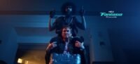 A man sits atop another mans shoulders. He is holding guns in each hand and duct taped to the man he is on top of. (Hold the Fort)