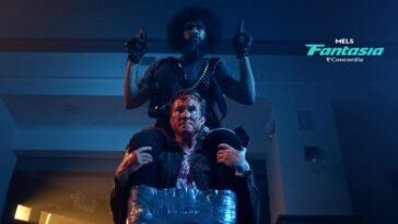 A man sits atop another mans shoulders. He is holding guns in each hand and duct taped to the man he is on top of. (Hold the Fort)