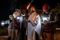 a group of masked individuals stand in a line, one is holding a torch (Self-Help)