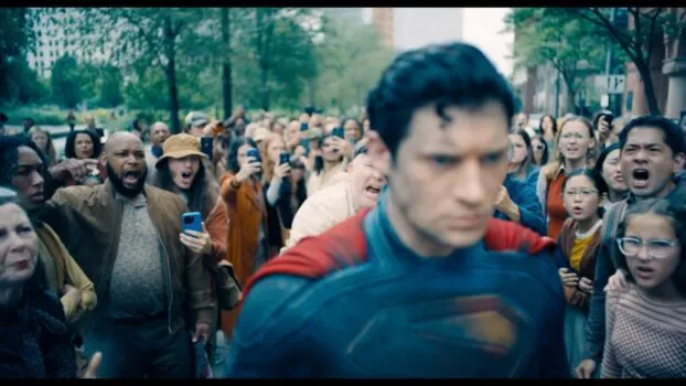 Superman walks through a crowd, with people yelling and screaming at him. A man throws a can at his head.